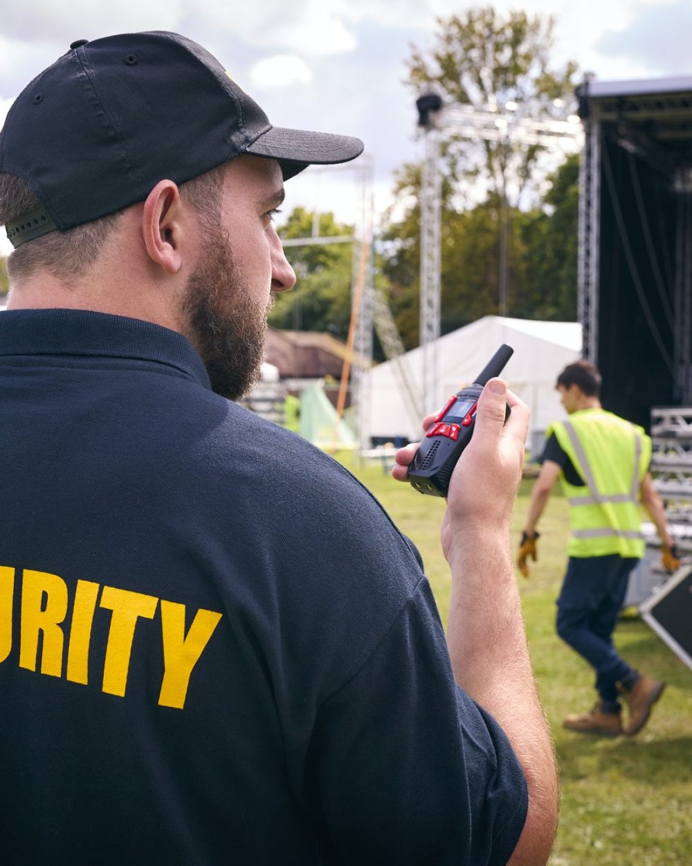 Rear View Of Security Team At Outdoor Stage For Music Festival Or Concert Talking Into Radio